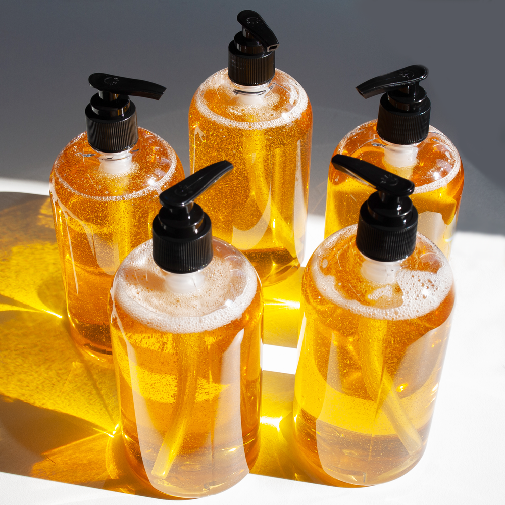 5 golden liquid soap bottles form a pentagon in front of a white and grey background. Sun light illuminating them